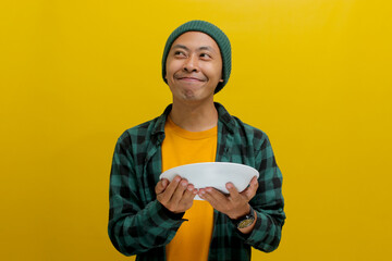 Hungry young Asian man looks towards an empty copy space while holding a plate in his hand,...