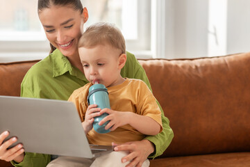 A woman is seated on a couch, engaging with a child who is actively using a laptop. The woman...