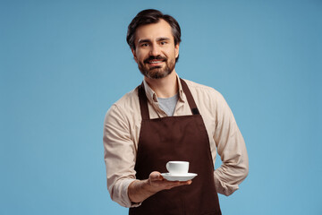 Smiling barista in brown apron holding cup and saucer in studio, isolated on blue