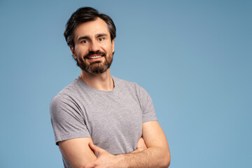 Smiling young bearded man with arms crossed looking at camera, posing isolated on blue