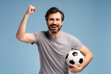 Happy excited bearded young man holding soccer ball and cheering, isolated on blue