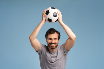 Portrait of happy young man holding football ball above head, looking at camera in studio