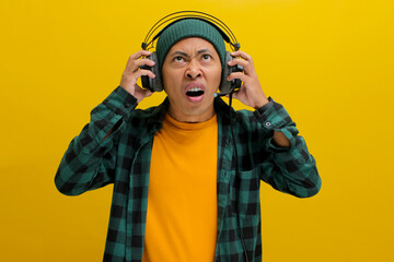 Annoyed Asian man in a beanie and casual clothes groans with frustration while wearing headphones....