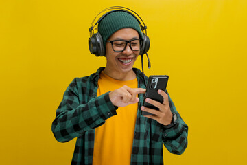 Asian man in a beanie and casual clothes browses a music app on his phone, selecting a song from a music app while wearing headphones. Isolated on a yellow background.