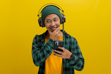 Young Asian man, deep in thought while listening to music on headphones. He holds his phone and gazes upwards, seemingly lost in the music or browsing the music app. Isolated on a yellow background.