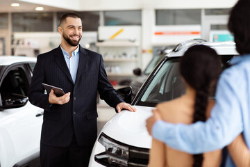 A professional salesman, clad in a smart suit, stands by a shiny new car with a digital tablet in...