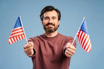 Positive bearded man celebrating and showing two national flags of United States on blue background