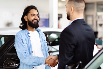 Two men in business attire are standing in front of a black car, engaging in a handshake. The...