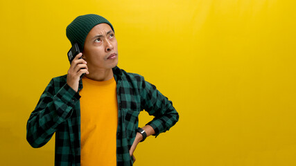 Serious young Asian man, dressed in beanie hat and casual shirt, listens intently to a serious conversation on his mobile phone, seemingly absorbing bad news, displaying worry, anxiety, and confusion