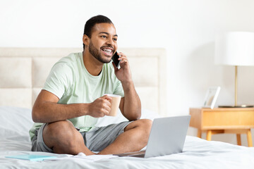 A cheerful African American man sits comfortably on his bed with a cup of coffee in hand, engaged...