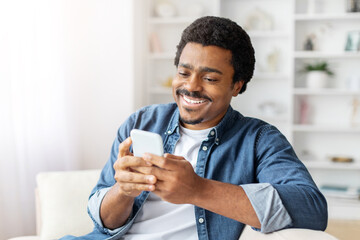 African American man is seated on a couch, engrossed in his cell phone, scrolling through content...