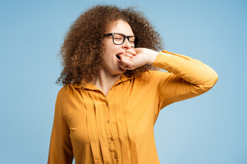 Tired young woman wearing glasses yawning, standing isolated on blue background. Morning concept