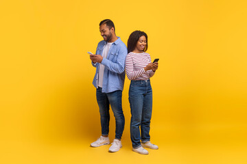 African American man and woman are standing with their backs touching, each absorbed in their own smartphone. They are casually dressed and appear to be enjoying a moment of connectivity