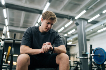 Low-angle portrait of fitness guy checking time of training in modern gym, looking at watch with satisfied expression and pleasant face, happy to finish exercise with barbell. Concept of healthy life.