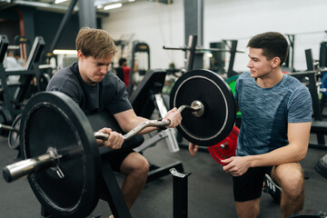 Portrait of motivated beginner sportsman hard time working out in gym with personal trainer at gym. Trainer helping beginner sportsman for correct doing Scott Curls with barbell, lifting up EZ-bar.