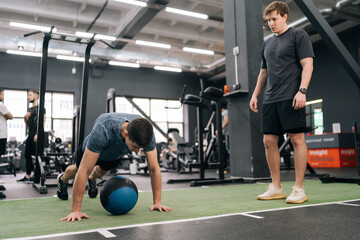 Professional sport coach explaining to beginner sportsman how to training with heavy fitness ball at gym. Motivated young man working out with medicine ball support under supervision of trainer.