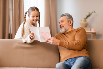 A young girl eagerly presents a hand-drawn heart card to her gray-haired grandfather as they share...