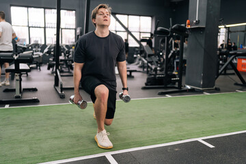 Portrait of motivated young beginner athletic sportsman in activewear holding dumbbells doing squat lunges warm up training indoor at gym. Concept of healthy lifestyle, physical sport activity.