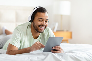 African American man is laying comfortably in bed, wearing headphones and engrossed in using a...