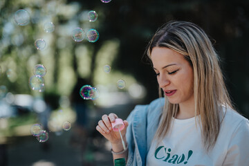 In a serene park setting, a young woman is captured blowing soap bubbles, radiating happiness and a carefree spirit under the sunshine.