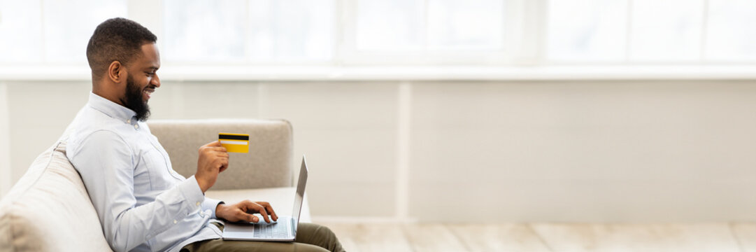 Easy Payment And Cashback Concept. Side view portrait of smiling african american young man holding credit card in hand and using laptop, sitting on couch at home, typing on keyboard, copy space