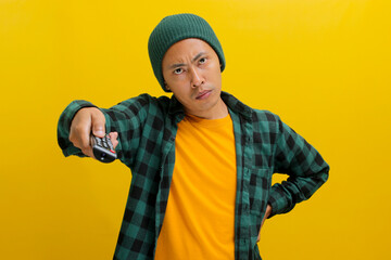 Bored young Asian man, dressed in a beanie hat and casual shirt, holds a TV remote control,...