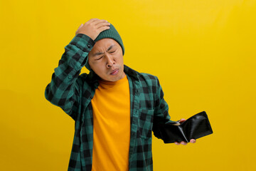 Broke Asian man dressed in a beanie hat and casual shirt, scratches his head in confusion as he...