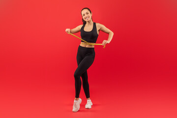 A cheerful young woman is standing against a vivid red backdrop, holding a measuring tape around her slim waist to showcase her fitness progress