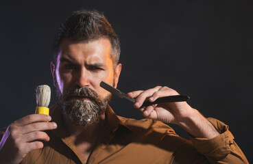 Handsome bearded man with straight razor and shaving brush. Barbershop accessories. Portrait of barber or hairdresser with beard and mustache with razor and brush for shaving. Professional beard care.