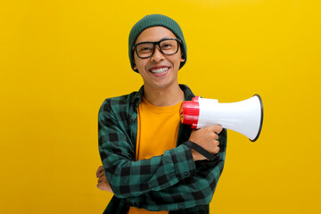 Confident Asian man in a beanie hat and casual shirt holding a megaphone, isolated on a yellow...