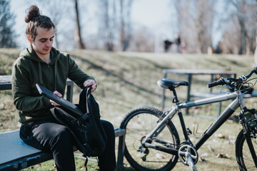 A young adult man relaxes in a park, unpacking his backpack while seated on a bench, with his...