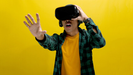 Excited Asian man wearing a VR headset reaches out with his hand, seemingly interacting with a virtual object in a VR game. Isolated on a yellow background