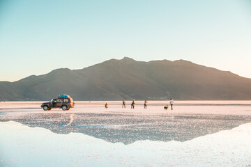 Fototapeta premium Reflections at Salar de Uyuni during sunset -April- - 4x4 Jeep and tourists behind mountain and mirror lake