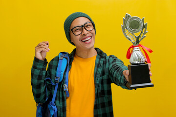 Happy young Asian student, adorned in a beanie hat, eyeglasses, and a casual shirt, proudly lifts...