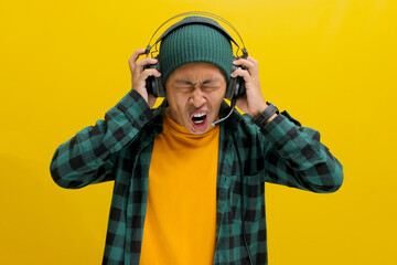 Annoyed Asian man in a beanie and casual clothes groans with frustration while wearing headphones....