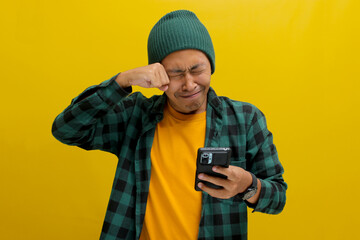 An unhappy Asian man, dressed in a beanie hat and casual shirt, holds a phone, visibly upset,...
