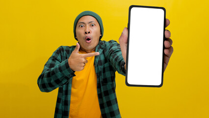 Excited Asian man, dressed in a beanie hat and casual shirt, excitedly showcases and points towards a mobile phone with an empty white screen, suggesting or recommending a new app or a special offer