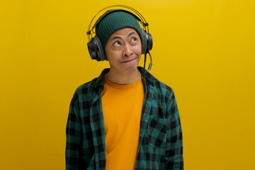 Young Asian man in a beanie and casual clothes, lost in thought while listening to music on his headphones. He gazes upward, a thoughtful smile playing on his lips. Isolated on a yellow background
