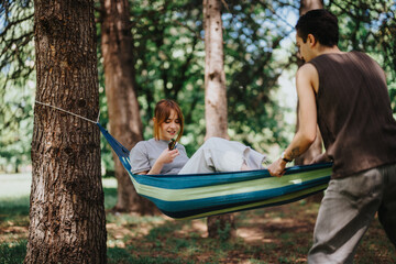 A serene outdoor setting features a young woman in a hammock reading a book, smiling at a man standing beside her in a park.