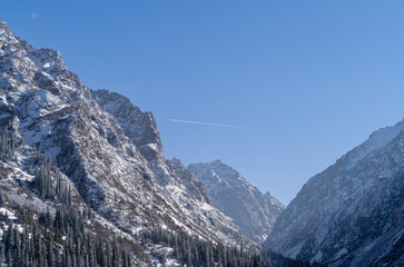 High flying plane in the blue sky, over a snow-covered canyon of towering rocky mountains, with a...