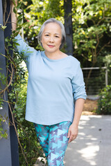 Outdoors, senior Asian female leaning on a post, wearing a light blue top