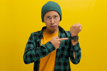 Frowning Asian man in a beanie and casual shirt impatiently taps his wristwatch, glancing at the...