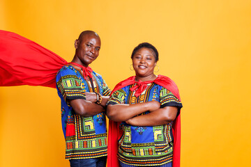 Obraz premium Smiling black couple dressed in superhero red cloaks posing with folded hands and looking at camera. Cheerful superman and superwoman pair wearing capes showing power and strength portrait