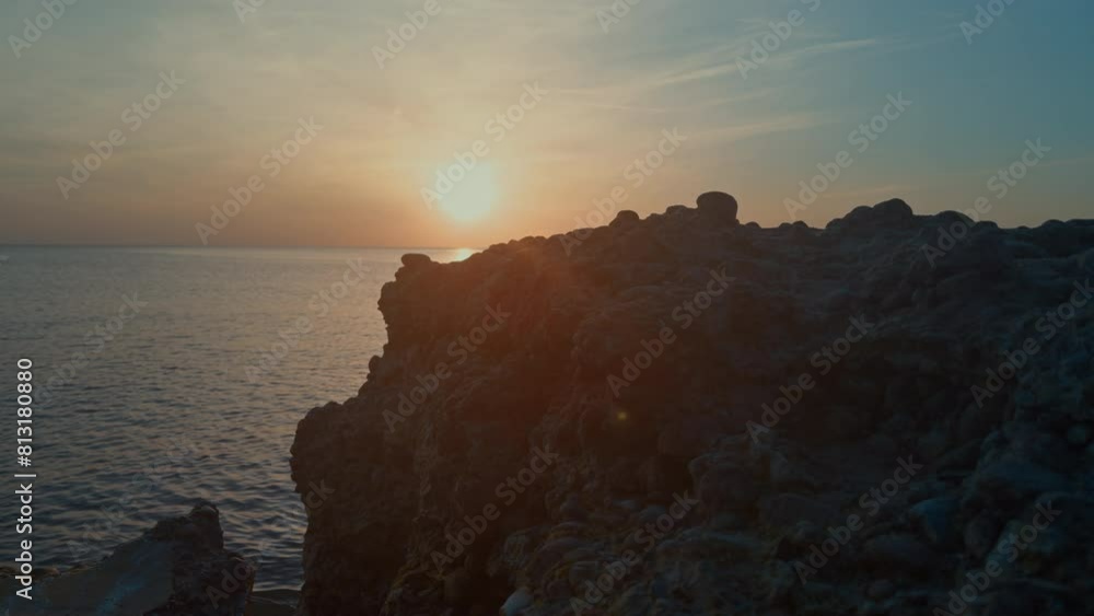 Wall mural a rocky cliff overlooking the ocean at sunset. the sun is setting and the sky is a mix of orange and