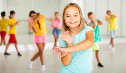 Smiling little girl training movements of modern vigorous dance with group of tweens in children...