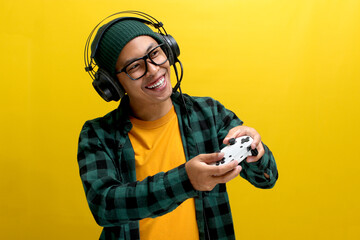 An enthusiastic Asian gamer, adorned in a beanie hat, casual shirt, and headphones, celebrates victory while playing a video game on a console with a gamepad or joystick isolated on yellow background.