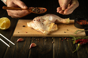 Close-up of a chef hands with a spoon adding pepper to a chicken leg. Broiler leg on cutting board...