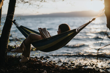 A carefree young girl loses herself in a book, swaying in a hammock beside a serene lake as the sun...