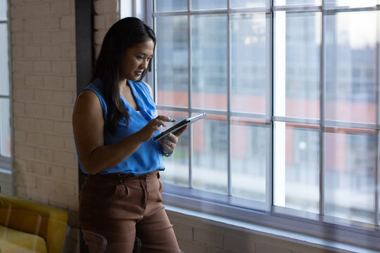 At office, biracial businesswoman wearing blue top, using tablet by window, copy space