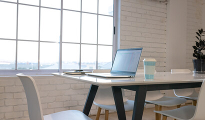 A laptop shows a spreadsheet on a white office table near a window with coffee